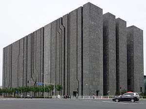 A dark gray block-like building towering over an intersection, with narrow, sometimes diagonal cracks in one side and large gaps in the other