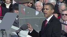 Barack Obama in a black coat gestures and talks at a lectern surrounded by teleprompters. Behind him, in the background, are about a dozen warmly dressed onlookers.