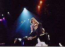 A blond woman riding a mechanical bull. She wears a T-shirt embedded with the flag of United States. Rays of light fall on her from above.