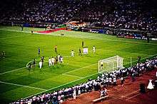 A wide-angle photograph of several football players preparing for a free-kick to be taken.