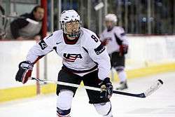 A female ice hockey player is skating towards the camera.  Her jersey is white and features a stylized "USA" on the front.