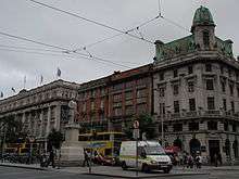 A busy street with large buildings, many cars and tram power cables overhead.