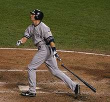 A man wearing a gray baseball uniform and navy-blue helmet drops his black baseball bat behind him after a swing.