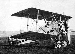 Three quarters view of military biplane on the ground with pilot in cockpit and check paint pattern on engine cowling and wheels