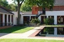  Two sides of a brick courtyard with white cloister behind a green lawn and a reflecting pool