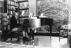 A black-and-white photograph of an elderly man with glasses sitting at a piano in front of a glass wall. Behind him is a full bookshelf.