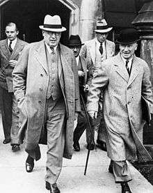 Two men in the foreground walk down a sidewalk, followed by three men in the background. The man in the left of the foreground is portly and wears a fedora, round-rimmed glasses, and a three piece suit. The man on the right is wearing an overcoat and a bowler hat, and holds a cane in his right hand and some documents in his left.