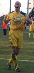 A bald man wearing yellow football shirt, shorts and socks and black football boots. He is smiling broadly and clenching his fists. He is standing on a grass surface and several people and indistinct structures are behind him.