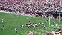 American football players line up in a goal line formation on an American football field surrounded by crowded stands.