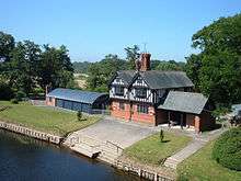 A small, two-storey, two-bay house seen from an elevated angle; the lower storey is in brick and the upper storey is timber-framed.  To the right of the house is a low extension and to the left is a barn with a curved roof.  In front of the house is a lawn and a slipway leading down to the river.