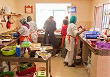 3 trainees dressed in white, cooking with the restaurant's chef who is dressed in black. A young volunteer is taking the food to serve to customers.