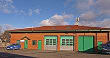 A large, sunlit, single-storey orange brick building with a roof covered with red tiles. On the left are two small green doors, in the centre are three large, green, shutter doors. Atop the roof is an aerial.