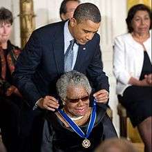 An elderly African-American woman, seated, smiling broadly, and dressed in black, being given an award by an African-American man in his fifties, wearing a blue tie and leaning over from behind her.