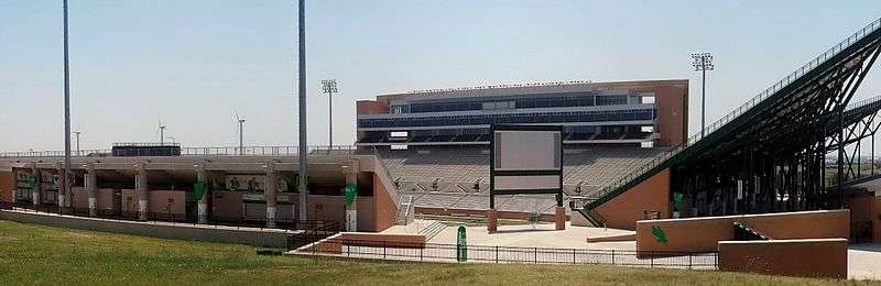 A wide photograph of an empty stadium. Three wind turbines are in the distance on the left, luxury boxes centered, and v-shaped stands are on the right.