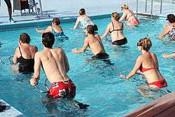 A group of people in swimwear pedaling on metal frames in a shallow pool