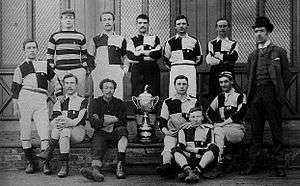 Eleven young men in sports clothing and one man wearing a suit pose for a team photo. One of the players is of Afro-Caribbean appearance.