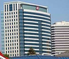 A white and blue building with a red logo on top stands behind and next to several short buildings.