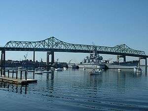 A large ship resting on the water under a bridge, smaller boats are visible in the foreground along with a ripple effect on the water.