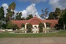 Photograph of the Botanic Building, one of several buildings in Balboa Park. A red conservatory building, it is fronted by a reflecting pool and surrounded by multiple species of trees.