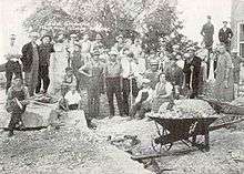 A wheelbarrow full of debris sits in the foreground, while townspeople and workers look at the unknown photographer, apparently taking a break from cleaning up debris after the bombing at the school