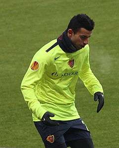 A photograph of a dark haired footballer who is seen in the process of training. The man is wearing a light green training shirt, black gloves, dark blue shorts and a dark blue scarf wrapped around his neck.