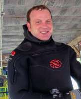 A smiling man with short brownish hair and ruddy cheeks smiling and wearing a dark blue wetsuit