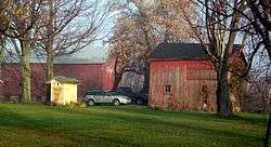 Two large red wooden buildings, the one on the left bigger than the other, and a small yellow one in front of it. seen through some trees in the foreground