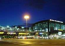 The exterior of a railway station at night. A rectangular building on the right has 2 of its sides visible, both of their walls windowed. A shorter connecting structure on the left is suspended over a street and lined with advertisements.