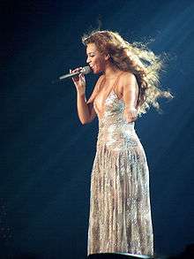 A black female singer with long, dark hair, singing on stage into a microphone in her right hand, wearing a silver, full-length dress.