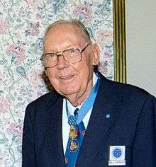 Elderly white man wearing a suit, tie, and glasses, with a medal hanging from a ribbon around his neck.