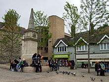 A paved square with a war memorial. People sitting on the steps. Pigeons waiting to be fed. Shops in the background.