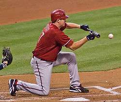 A man in a red baseball jersey and cap and gray baseball pants kneels on a dirt and grass field. He is holding a baseball bat flat in front of him in his hands with a ball approaching it.