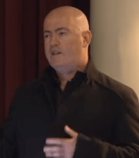 A close-up image of bald man, who looking up behind the camera while talking at a conference.