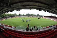 The inside of a football ground as photographed from one of the touchlines. Red seating is visible in the foreground, with the pitch in the middle and the opposite touchline in the background. A standing-only area is visible to the right of the picture. Football players and officials can be seen on the pitch.