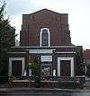 A plain building in very dark brick. In the foreground is a flat-roofed entrance area with two wooden doors and a small window (partly obscured).  Behind this, a squat tower, with a central window,  rises in several stages.