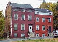 Two red three-story-tall brick buildings, with a small attic level and side-gabled roofs, on a street that slopes down to the right of the image