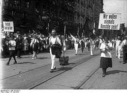 People holding banners and waving flags march down a street. Lining the road are crowds of supporting people.