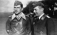 Black-and-white photograph showing half-length view of two uniformed men outdoors, standing next to each other. The young man on the left is wearing a field cap and a pilot's leather jacket with fur collar, with an Iron Cross displayed at the front of his shirt collar. The middle-aged man on the right is shown in three-quarter profile; he is smiling and wears a jacket with military decorations.