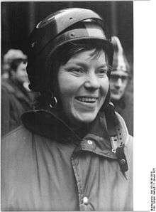 A smiling brunette woman wearing a winter jacket and a full-face helmet with a lifted visor.