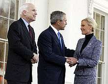 White-haired man in dark suit looks on as grey-haired man in dark suit holds hand and greets blonde-haired woman in medium-colored suit, all in front of a white building.