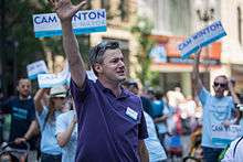 A man in his mid-30s wearing a purple polo shirt waves as people behind him hold up signs with his name, Cam Winton.