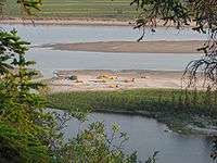 Canoes and tents rest on a sandy spit along a river.