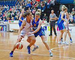 A female baseball player is attempting to drive to a basket while another female player is guarding her, and attempting to reach for the ball.