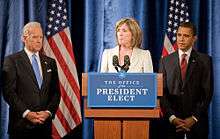 A pale-skinned man in his sixties with white hair, a pale-skinned woman in her fifties with light brown hair, and a brown-skinned man in his forties with dark hair, all stand in front of a blue drape backdrop with two American flags. Both men are wearing dark business suits with white shirts and ties, one blue, one red; the woman is wearing a light beige suit jacket. The woman is behind a brown podium with a blue and white sign saying "Office of the President Elect" and two black microphones. All three are looking serious and both of the men have their hands folded in front of them.