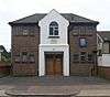 A two-storey building with a dark brick façade dominated by a central gabled stuccoed section. This has recess for the wooden entrance door and an arched window.  Both storeys have six small windows with blue frames.