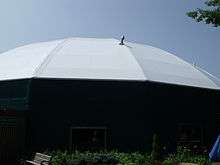 A round, green building with a white tent-like roof stands against a clear summer sky.