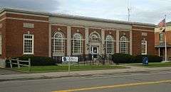 A one-story brick building with elaborate classically-inspired stone detailing, seen from across a street. There are five round-arched windows grouped in the center and two rectangular ones nearer the ends. At the right is an American flag flying from a pole and a mailbox.