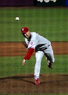 A man in a gray baseball uniform with red sleeves and cap throws a baseball with his left arm.