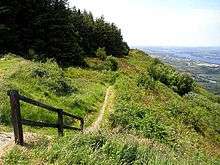 Dirt footpath skirting the edge of a steep grassy descent, with trees on the left and a distant countryside view below to the right.