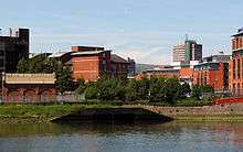 The open end of a river culvert in the bank of a much wider river, with office buildings and development visible above the waterside.